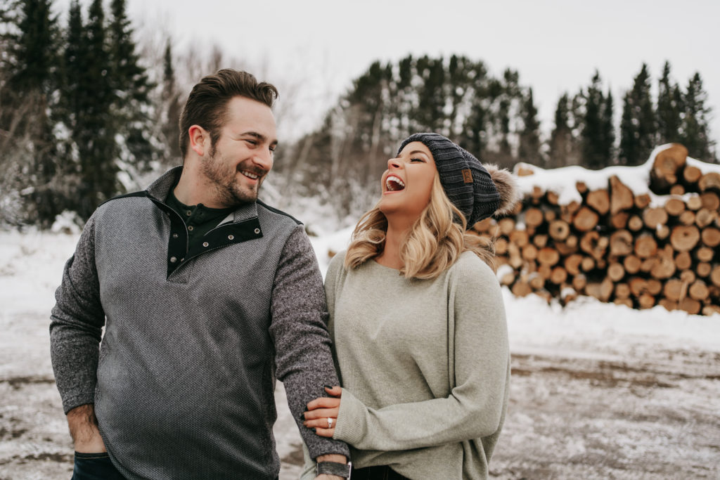 An engaged couple laughs in front of a wood pile in Northern Minnesota during their engagement session.