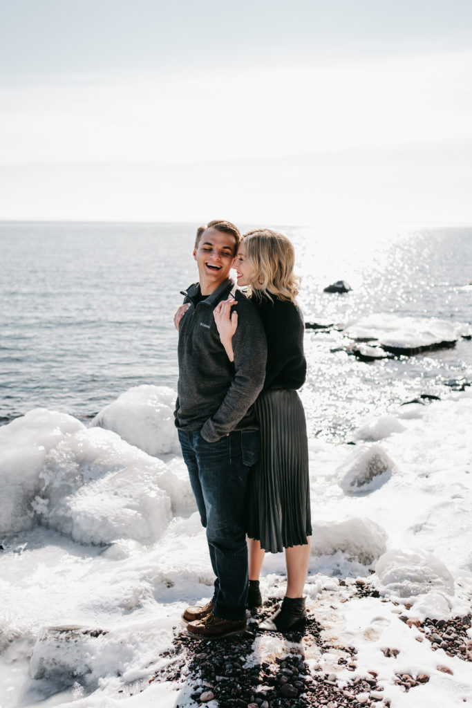 An engaged couple laughs near Lake Superior during their winter engagement session