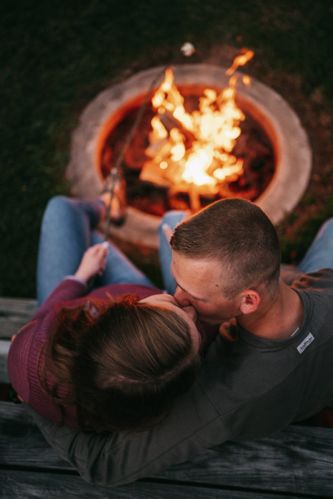 An engaged couple kisses by a campfire.