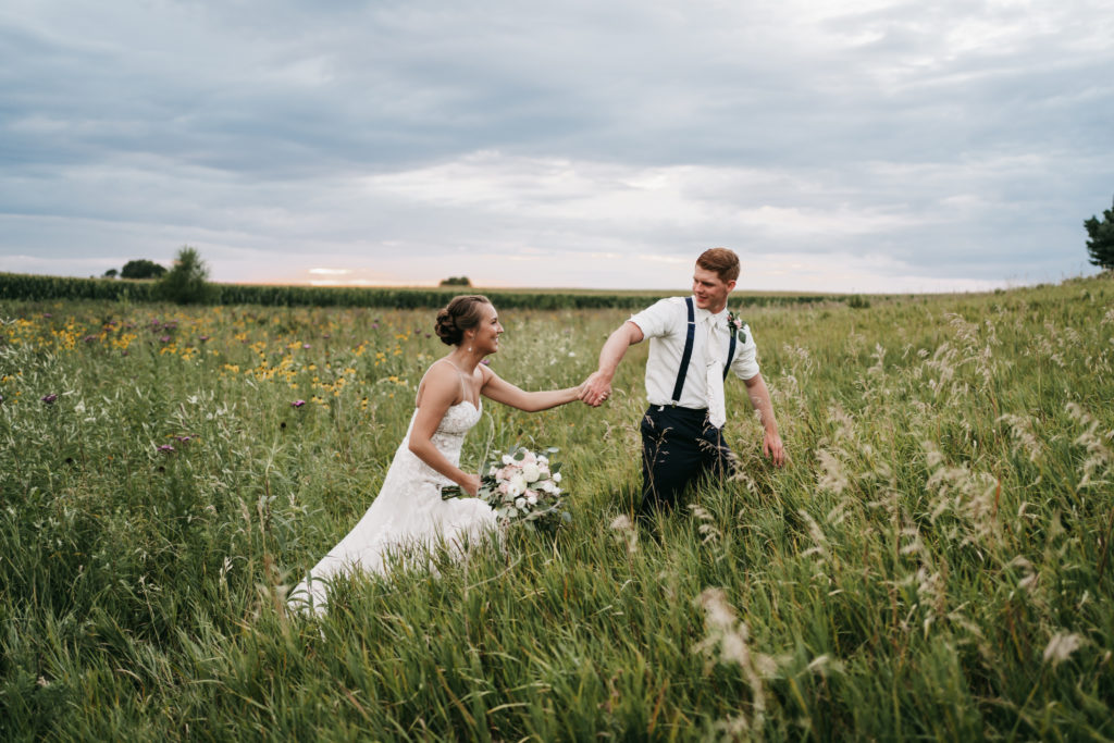 Bride and groom hold hands while walking through an open field at sunset.