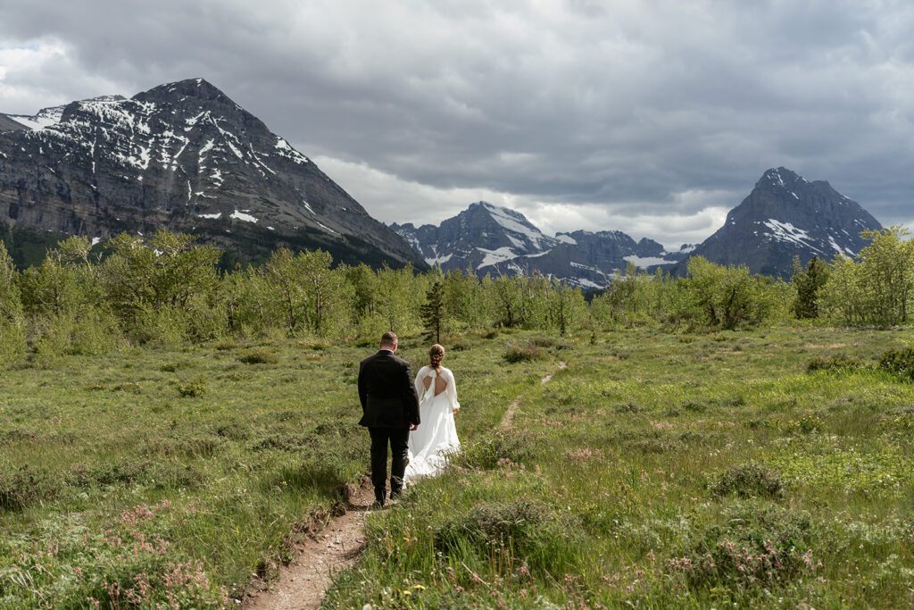 Bride and groom stroll down a dirt path in the Many Glacier District in Glacier National Park with an overcast sky ahead, and greenery surrounding them.