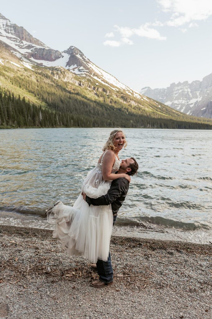 A groom playfully lifts his bride up on the shore of Many Glacier during an intimate wedding photographed by Sydney Breann Photography.