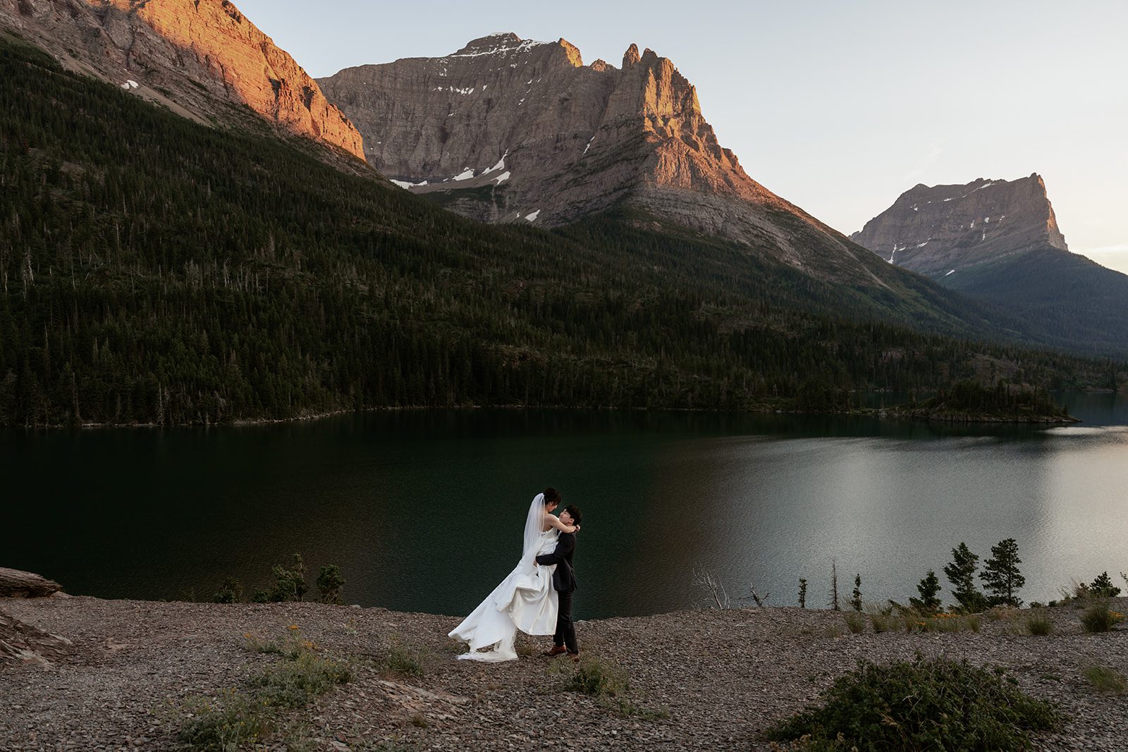 A warm alpenglow paints the mountains at Glacier National Park, where a bride and groom playful spin around atop Sun Point, overlooking St. Mary Lake.