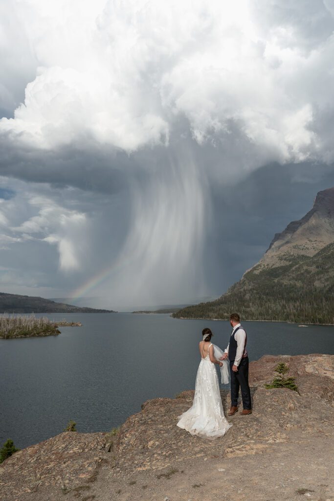 Bride and groom take in the beauty of storm clouds and a rainbow over Sun Point in Glacier National Park