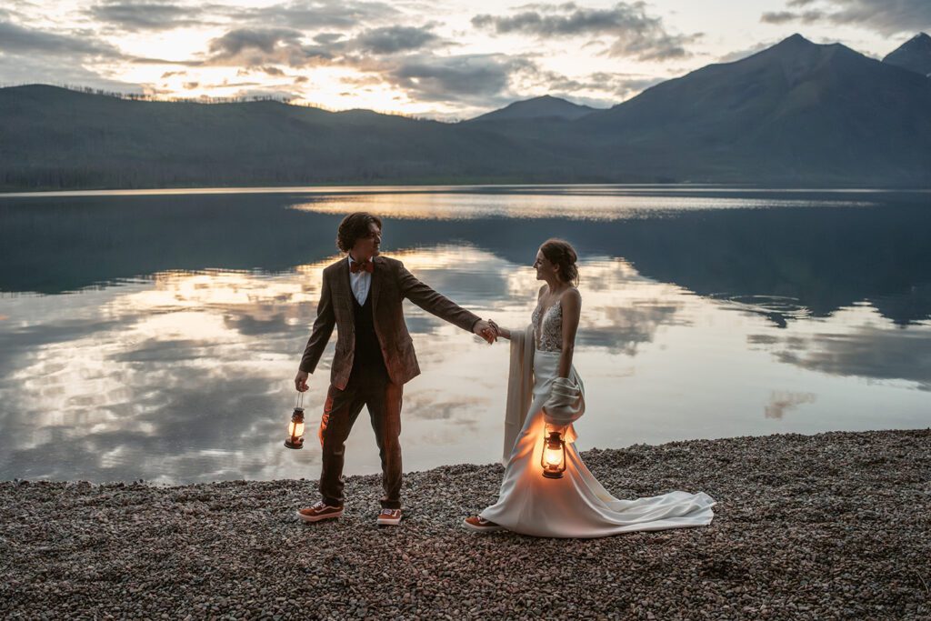 Newlyweds walk hand-in-hand along the shore of Lake McDonald with lanterns during blue hour in Glacier National Park.